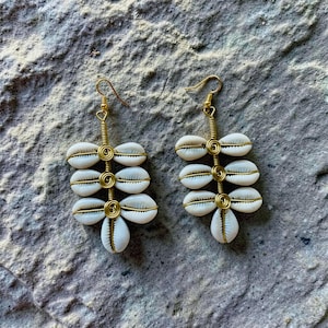 May include: A pair of handmade earrings featuring white cowrie shells arranged in a cascading design. The shells are connected with gold-colored wire, creating a unique and natural aesthetic. The earrings have gold-colored ear wires.
