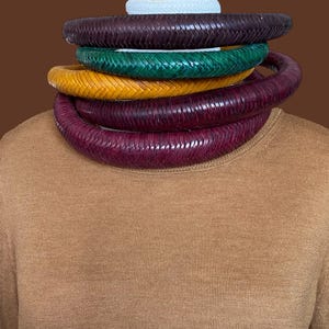 May include: A stack of five woven leather necklaces in various colors: dark purple, green, yellow, and burgundy. The necklaces are stacked on top of each other, with a white woven band at the top. The background is a solid brown.