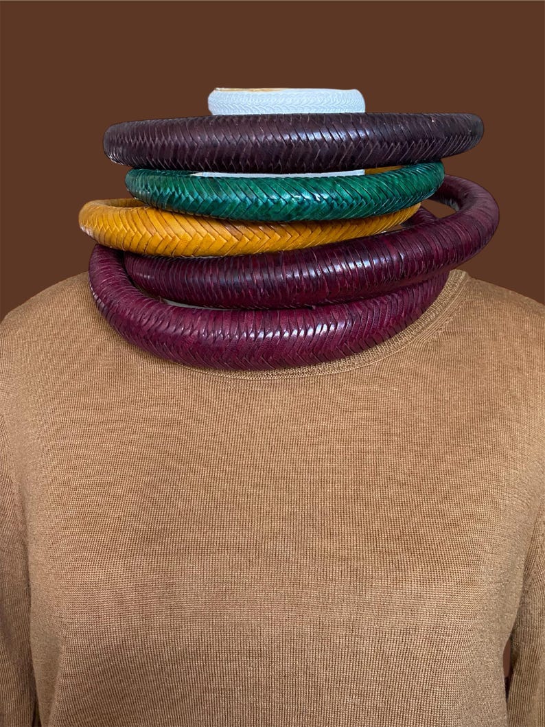 May include: A stack of five woven leather necklaces in various colors: dark purple, green, yellow, and burgundy. The necklaces are stacked on top of each other, with a white woven band at the top. The background is a solid brown.