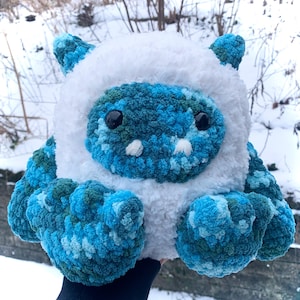 May include: A crocheted plush toy in the shape of a creature with a white fur hood, blue and green camouflage body, and black eyes.