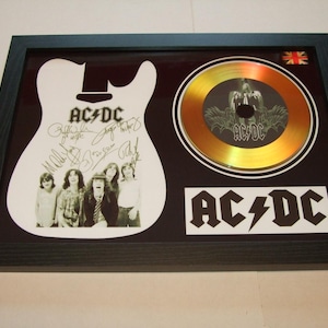 Puede incluir: Recuerdos enmarcados de AC/DC con un recorte blanco en forma de guitarra con las firmas de la banda y una foto en blanco y negro. Incluye un CD dorado y el logotipo de la banda. Se ve una pequeña bandera de la Union Jack.