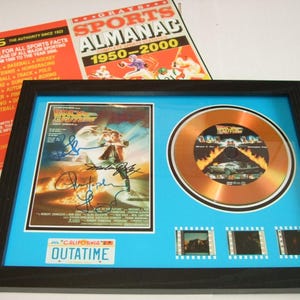 Puede incluir: Enmarcado recuerdo de Regreso al Futuro. Incluye un póster de la película firmado, un disco dorado, tiras de película y una matrícula de California que dice "OUTATIME". El marco negro tiene un fondo azul claro, con un Grays Sports Almanac visible.