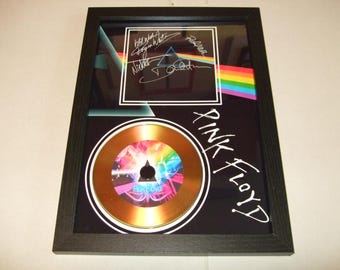 Może przedstawiać: Oprawiona grafika albumu Pink Floyd z płytą CD, logo zespołu i podpisami. Grafika zawiera pryzmat załamujący światło w tęczę, z napisem "Pink Floyd" w białym kolorze. Czarna ramka.