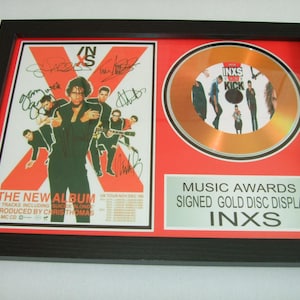 May include: Framed INXS music award display featuring a signed gold disc and a poster for "The New Album." The poster has band members and the text "INXS" in a bold, stylized design. The frame is black.