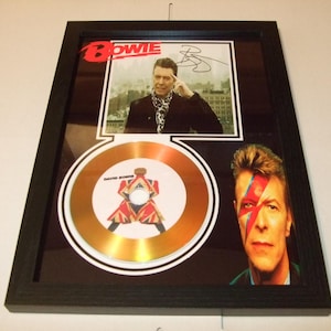 May include: Framed artwork featuring David Bowie. Includes a photo with a signature, a gold-colored record, and a portrait with iconic face paint. The word "BOWIE" is displayed in red. The frame is black.