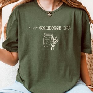 May include: A green t-shirt with the text "In My Sourdough Era" and a drawing of a jar with a wheat stalk.