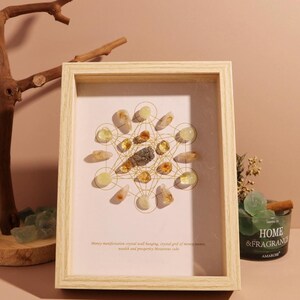 May include: A wooden frame with a white background and a crystal grid with gold wire. The crystals are various shades of yellow and green. The text "Money manifestation crystal grid hanging, crystal grid of money power, wealth and prosperity. Metaphysical gift." is printed below the grid.