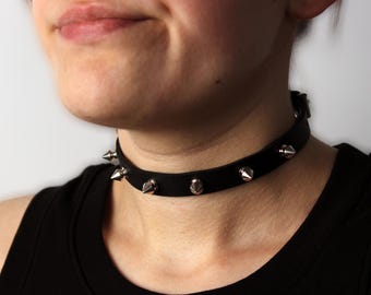 May include: A black leather choker with silver pyramid-shaped studs. The choker is worn by a person with a black shirt.