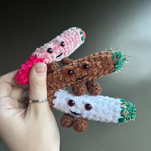 Pode incluir: Três brinquedos de pelúcia de crochê em forma de juntas, um rosa, um marrom e um branco. Cada brinquedo tem um rosto sorridente com olhos vermelhos e detalhes verdes.