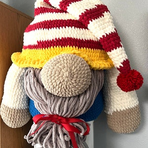 May include: A crocheted gnome with a red, white, and yellow striped hat. The gnome has a long, gray beard and a brown nose.