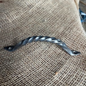 Peut inclure: Une poignée en métal torsadé, fabriquée à la main, avec une finition gris foncé. La poignée présente un motif texturé en forme de corde et est fixée avec de petits rivets sombres à chaque extrémité. Conçu pour un meuble.