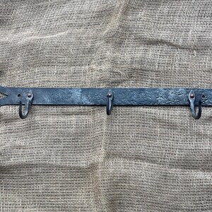 May include: Hand-forged metal wall hook with four hooks. The dark gray metal has a textured surface and curved ends. The hooks are evenly spaced along the horizontal bar, perfect for hanging coats, hats, or other items. The rustic design adds a touch of vintage charm.