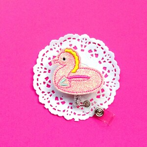 May include: Pink glitter flamingo pool float badge reel with a white lace background.