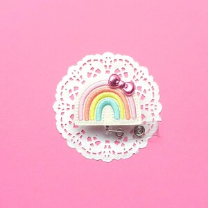 May include: A colorful rainbow badge with a pink bow, set on a white lace doily against a pink background. The rainbow features pastel colors: pink, yellow, blue, and green. The badge has a retractable reel.