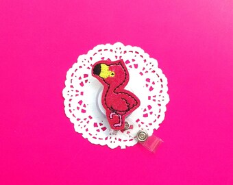 May include: A pink flamingo with a yellow beak and black eyes is attached to a white doily. The flamingo is on a retractable badge holder.