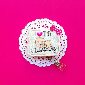 May include: White fabric badge with pink heart and embroidered text "I love tiny humans" and a cartoon bear. The badge is attached to a white doily and a clear plastic retractable badge holder.