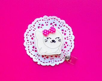 May include: White felt seal with pink polka dot bow, embroidered eyes and a smile. The seal is attached to a white doily and a clear plastic badge reel.