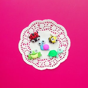 May include: Five colorful plastic animal figurines on a white doily. The animals include a ladybug, a snail, a bee, a frog, and a turtle.