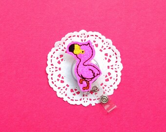 May include: A pink flamingo-shaped retractable badge reel with a glittery finish. The flamingo has a black beak, yellow eye, and is set on a white lace doily against a bright pink background. The reel has a clear plastic strap and a metal clip.