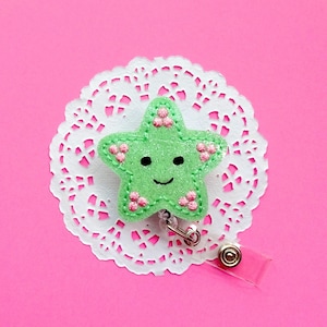 May include: A green felt star with a smiling face and pink flower accents is attached to a white lace doily. The star is attached to a clear plastic badge holder with a silver clip.