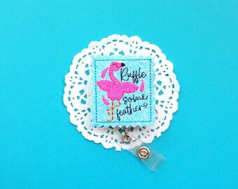 May include: A pink flamingo with the text "Ruffle some feathers" on a blue background. The badge is attached to a clear retractable badge holder.