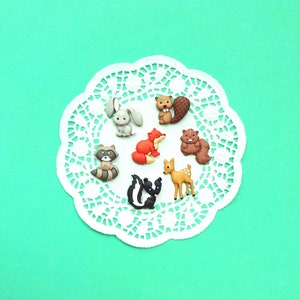 May include: A set of eight colorful animal-shaped buttons, including a fox, a deer, a raccoon, a beaver, a skunk, and a rabbit. The buttons are arranged on a white doily against a turquoise background.