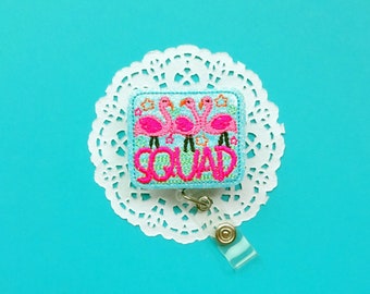 May include: A blue and pink embroidered badge with three pink flamingos and the word "Squad" on a white doily background. The badge is attached to a clear plastic retractable badge holder.