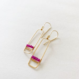 May include: A pair of gold-colored rectangular drop earrings. Each earring features a row of small, bright pink beads wrapped with gold wire. The earrings have a simple, elegant design with a hook closure.