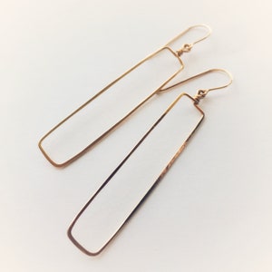May include: A pair of gold-toned rectangle shaped dangle earrings. The earrings are made of thin metal wire and have a simple, minimalist design.