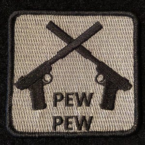 Puede incluir: Un parche bordado gris y negro con dos pistolas cruzadas y el texto "PEW PEW".