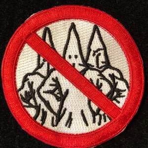 May include: A white embroidered patch with a red circle and a diagonal red line through it. The patch depicts two figures wearing pointed white hoods. The text "No" is written in black on the red line.