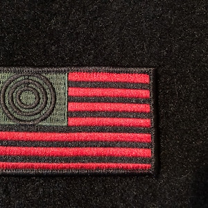 Può includere: Una toppa ricamata nera e rossa con un cerchio verde nell'angolo in alto a sinistra. Il cerchio ha un design a cerchi concentrici. Le strisce rosse e nere assomigliano a una bandiera americana.