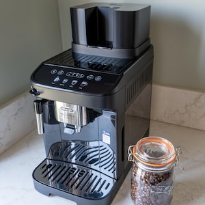May include: A black De'Longhi Magnifica Evo espresso machine with a stainless steel front panel. A glass jar filled with coffee beans sits beside the machine. The machine has a water reservoir and a drip tray.