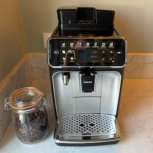 Puede incluir: Una máquina de espresso negra y plateada con una pantalla digital que muestra varias opciones de café. La pantalla muestra "My Coffee" y tiene iconos para diferentes tipos de café. La máquina tiene una bandeja de goteo y un tanque de agua.