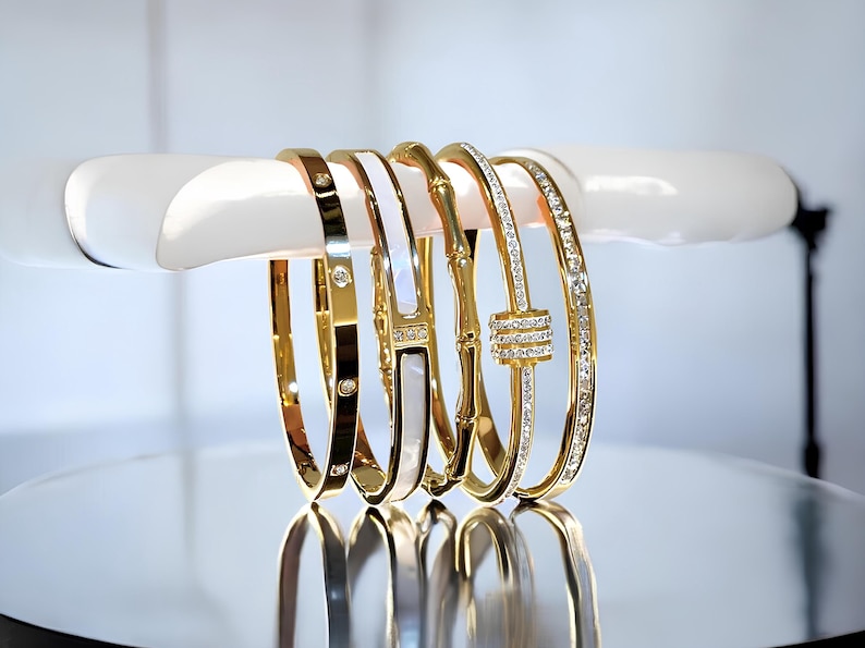May include: Four gold bracelets with different designs. One bracelet has a white enamel band with a diamond pattern. Another bracelet has a diamond-encrusted band with a knot design. The other two bracelets have a plain gold band with diamonds.