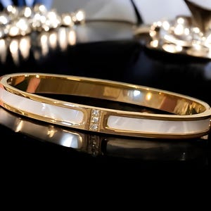 May include: A gold bangle bracelet with a white mother-of-pearl inlay and clear crystal accents.