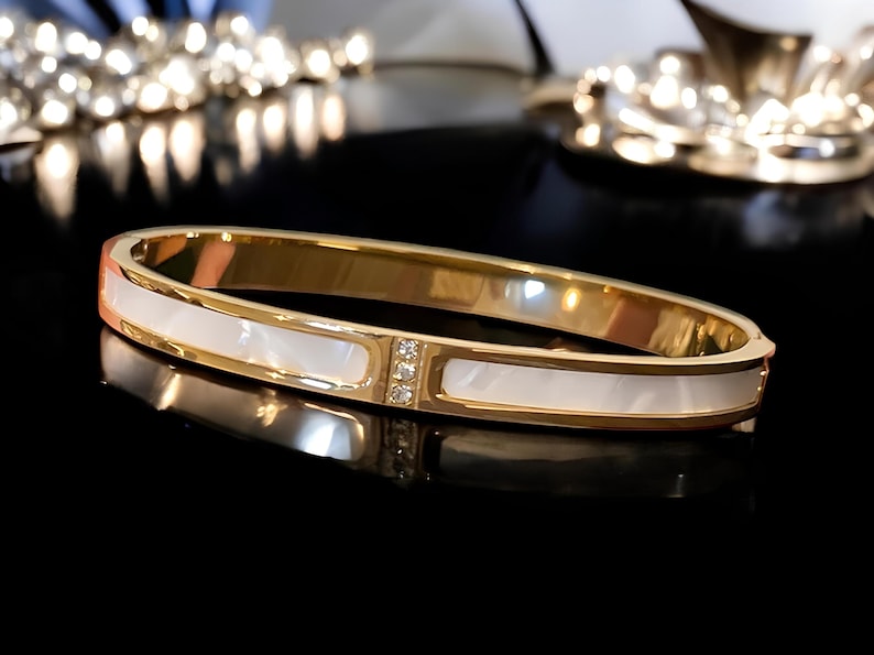 May include: A gold bangle bracelet with a white mother-of-pearl inlay and clear crystal accents.
