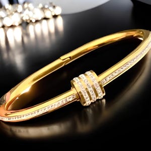 May include: A gold bangle bracelet with a decorative, round, crystal-encrusted center piece. The bracelet is made of metal and has a polished finish.