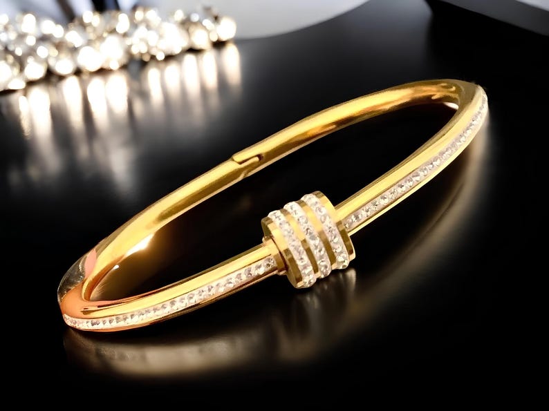 May include: A gold bangle bracelet with a decorative, round, crystal-encrusted center piece. The bracelet is made of metal and has a polished finish.