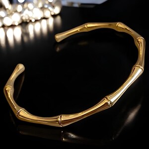 May include: A gold-toned bangle bracelet with a bamboo design. The bracelet is open and has a smooth, polished finish.