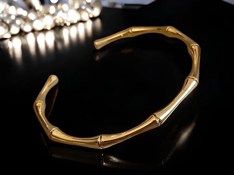 May include: A gold-toned bangle bracelet with a bamboo design. The bracelet is open and has a smooth, polished finish.