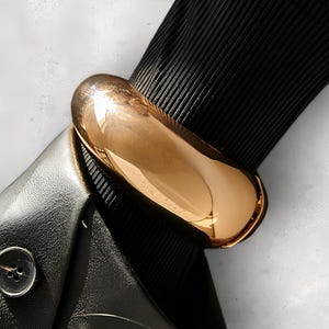 Peut inclure: Un bracelet jonc doré à la forme ronde et lisse. Le bracelet est porté sur un fond en tissu noir.