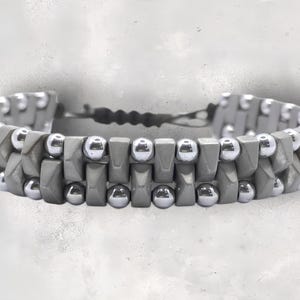 May include: A gray and silver beaded bracelet with a geometric design. The bracelet is made of square and round beads.