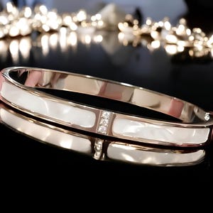 May include: A gold bangle bracelet with a white mother-of-pearl inlay and clear crystal accents.