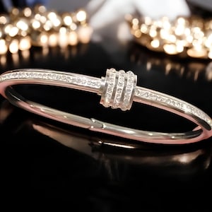 May include: A silver bangle bracelet with a row of clear cubic zirconia stones around the band and a larger cluster of cubic zirconia stones in the center.