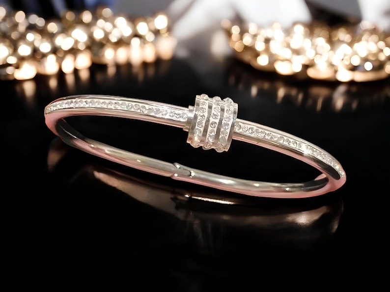 May include: A silver bangle bracelet with a row of clear cubic zirconia stones around the band and a larger cluster of cubic zirconia stones in the center.