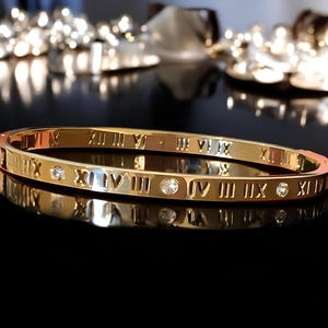 May include: A gold bangle bracelet with Roman numeral markings and small clear stones.