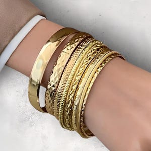 Op de afbeelding: Een set gouden armbanden met verschillende texturen en patronen. De armbanden zijn gestapeld op de pols van een persoon.