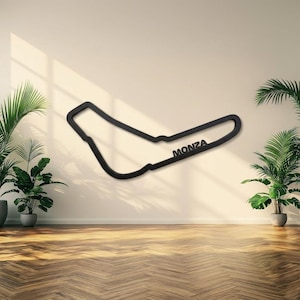 Può includere: Una scultura da parete nera che raffigura il profilo del circuito di Monza. L'oggetto presenta un design a cornice vuota e continua, con la parola MONZA integrata nella sezione inferiore del tracciato in lettere maiuscole e in grassetto.