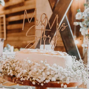 Könnte beinhalten: Eine weiße Hochzeitstorte mit einem hölzernen Tortendekor, auf dem "Mr & Mrs Story" steht. Die Torte ist mit weißen Gipskrautblüten verziert.